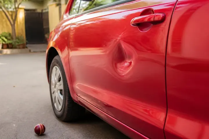 dent meaning picture: A close-up of a shallow hollow in a shiny red car door, a cricket ball lying on the road below.